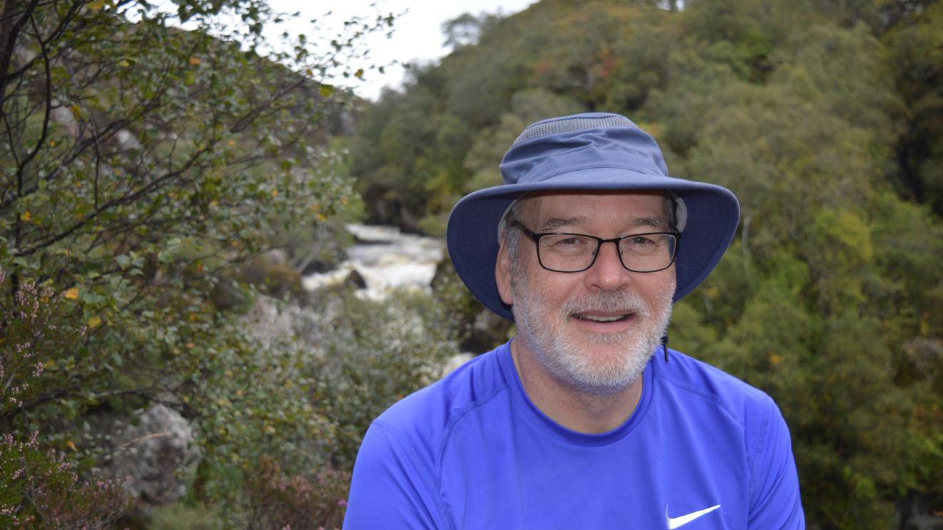 Neil, volunteer independent visitor standing in front of a watercourse with a blue bucket hat and wearing a blue t-shirt