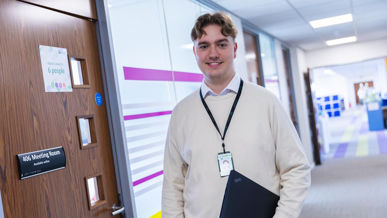 One our apprentices holding a laptop and standing outside a meeting room