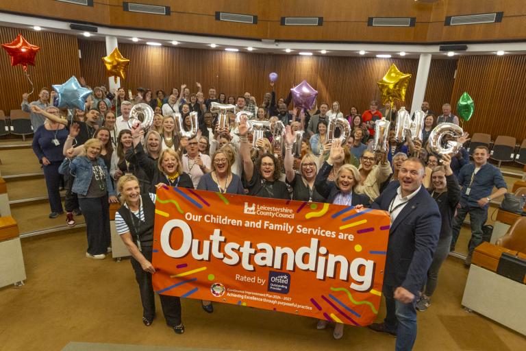 A room full of staff hold a large orange banner saying Leicestershire County Council Children and Family Services are outstanding