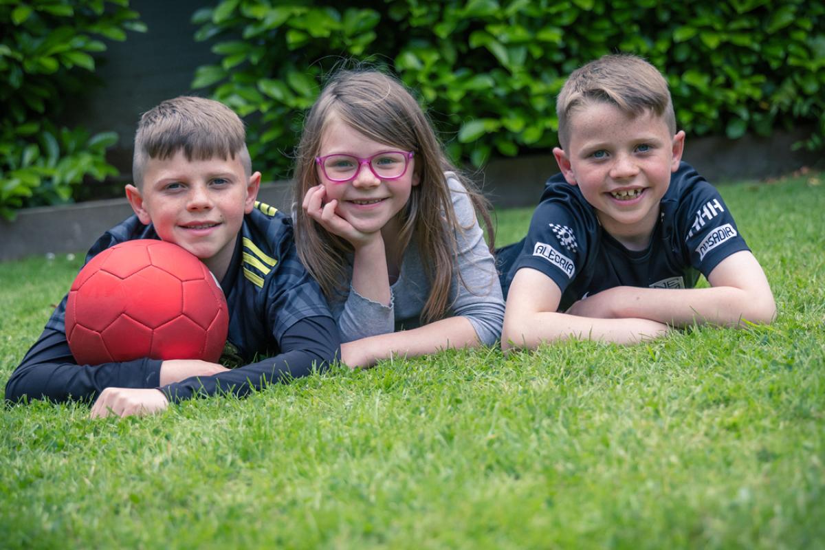 3 children on a grass lawn smiling, one with a red football